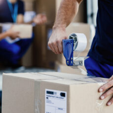 Close-up of delivery man closing carboard box with a tape while preparing packages for shipment.